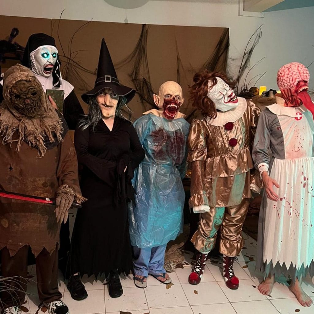 Group of people in elaborate horror costumes (nun, scarecrow, witch, zombie, clown, nurse) posing for a photo at a themed event