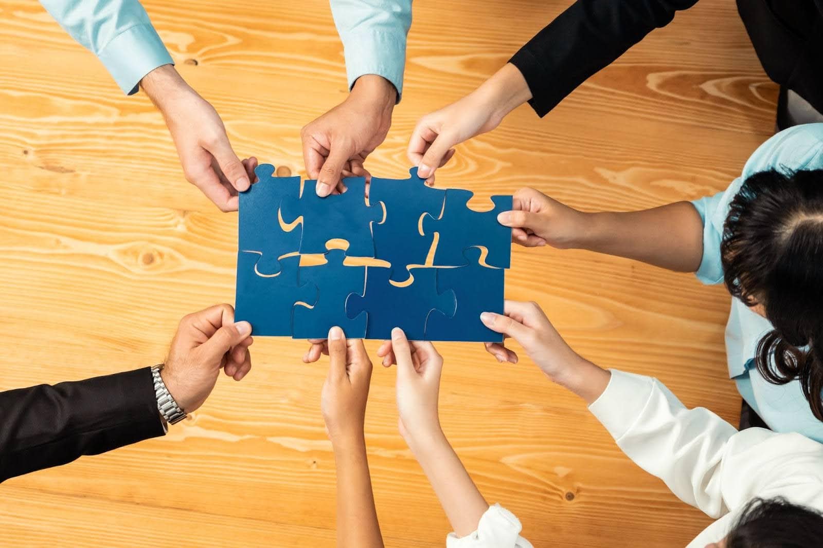An overhead shot of six sets of diverse hands working together to assemble a large blue jigsaw puzzle on a light wood-grain table, symbolizing teamwork and collaboration for building a corporate event team.