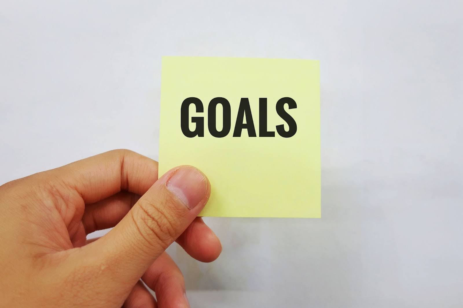 A hand holding a yellow sticky note with the word "GOALS" printed in bold, black, all-caps letters, emphasizing the importance of establishing clear goals and objectives for an event.