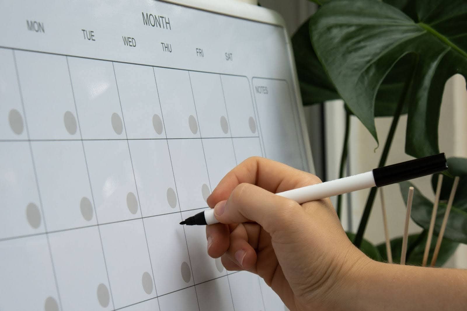 A close-up shot of a hand with a black marker poised over a blank monthly whiteboard calendar, symbolizing the detailed planning of logistics, operations, and risk management.