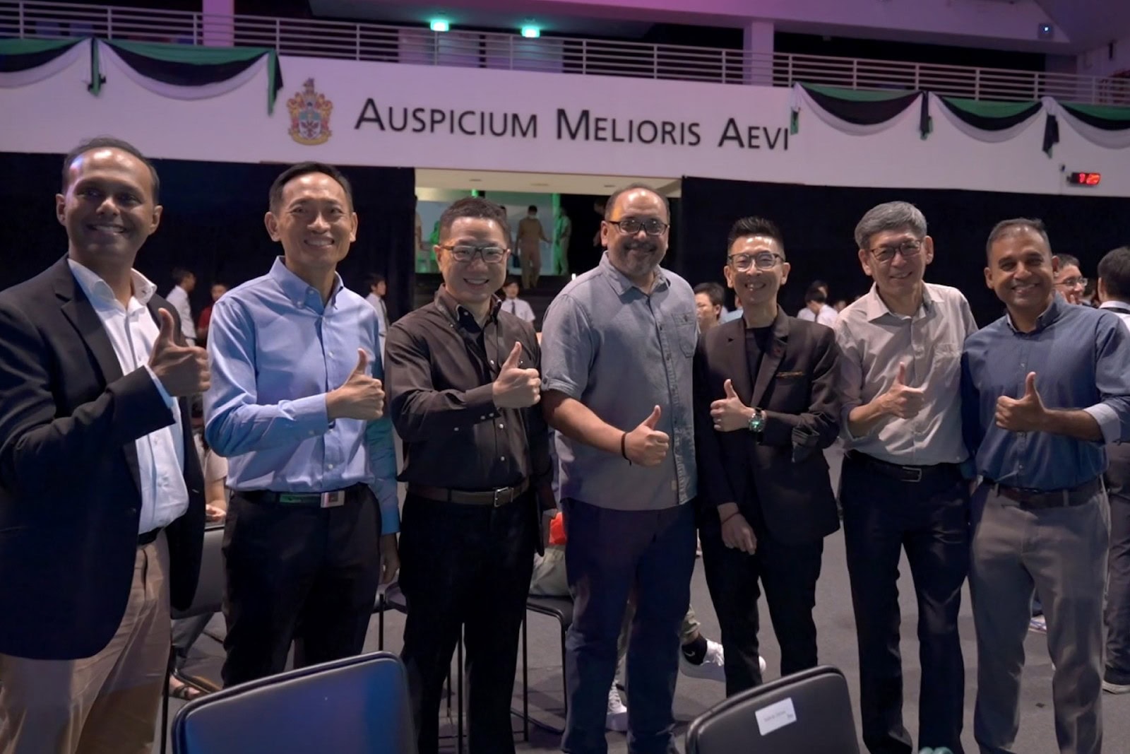 A group of seven smiling men in business and smart casual wear stand together giving the thumbs-up sign after a successful event, signifying a positive post-event wrap-up.
