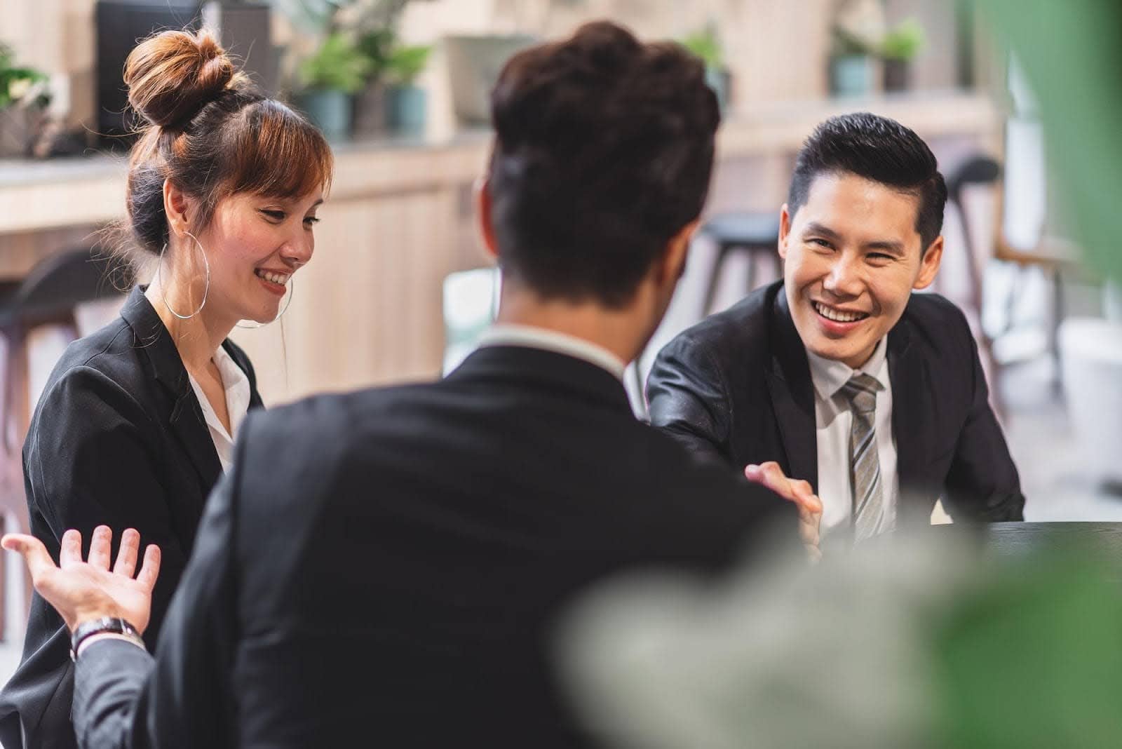 Three colleagues in business attire having a positive and collaborative meeting; a woman with her hair in a bun and a smiling man are actively engaged in a discussion with a third person, illustrating a key step in a corporate event planning guide.