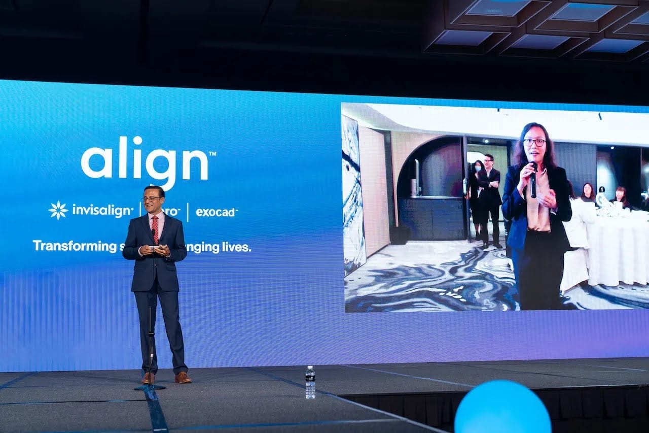 An example of corporate event planning: a man in a suit speaks on a professionally lit stage. Behind him, a large screen displays the 'Align' logo and a live video feed of a female speaker at a remote location, showcasing a modern hybrid corporate event.