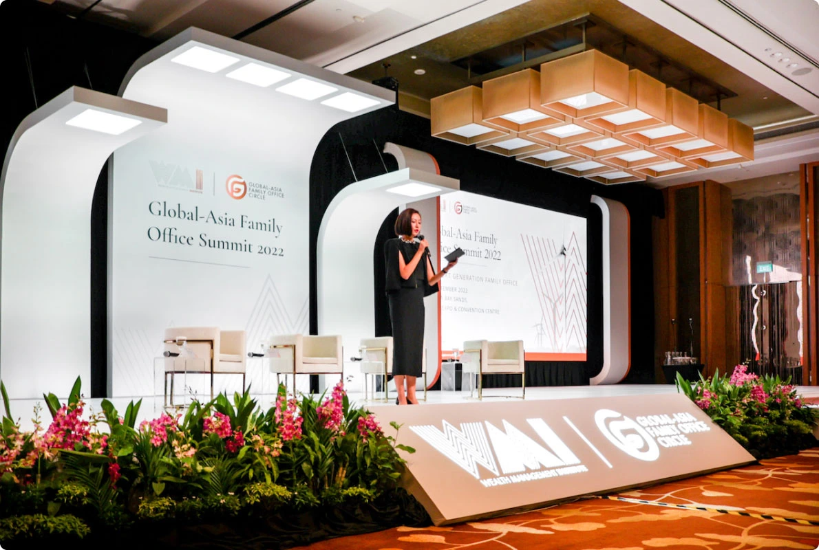 Corporate event speaker presenting on stage at the Global-Asia Family Office Summit 2022, with branded backdrop and floral arrangements.