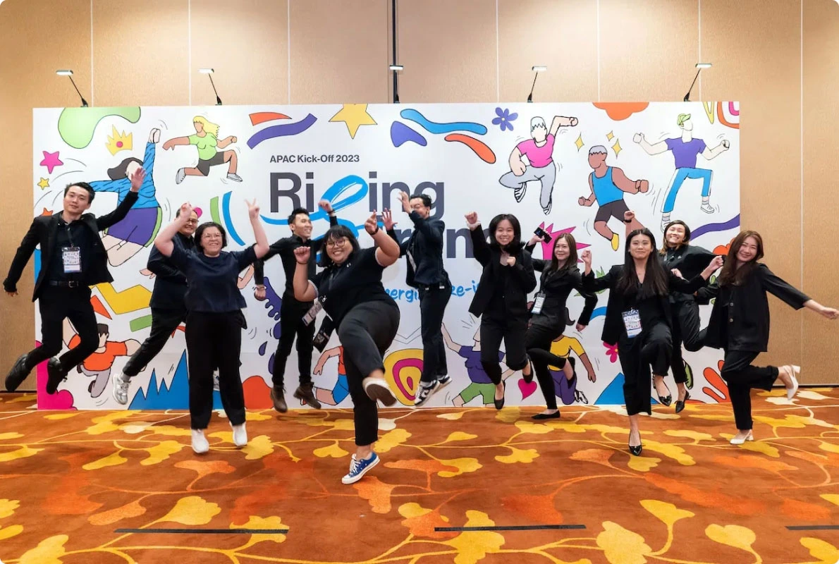 Event staffing team in black uniforms jumping and posing energetically in front of a colorful APAC Kick-Off 2023 backdrop at a corporate event.