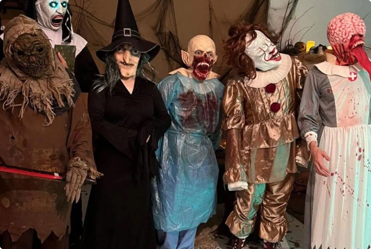 Group of performers in horror costumes at an experiential event, dressed as a witch, zombie doctor, evil clown, and other scary characters with detailed latex masks and special effects makeup.