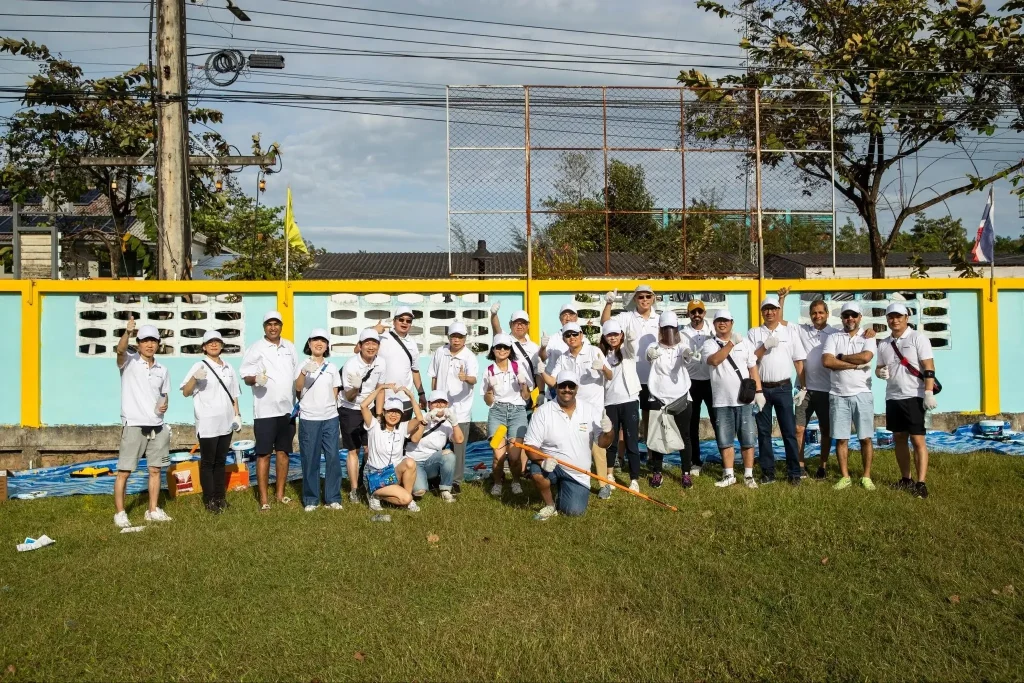 A group of volunteers pose cheerfully in front of a freshly painted mint-green and yellow wall holding paint rollers and brushes during a community service painting event outdoors.